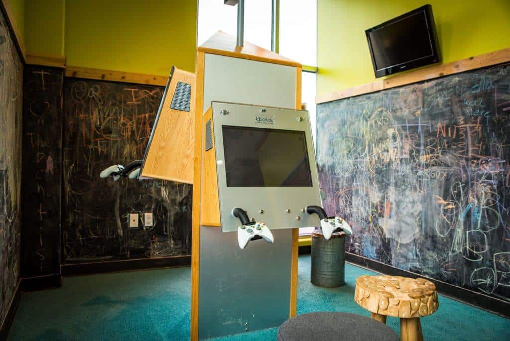 Kids play room with game systems in the center and huge blackboards covering the walls all the way to the floor, TV screen on the top right above one of the blackboards, blue wall to wall carpet and 3 legged stool type chairs