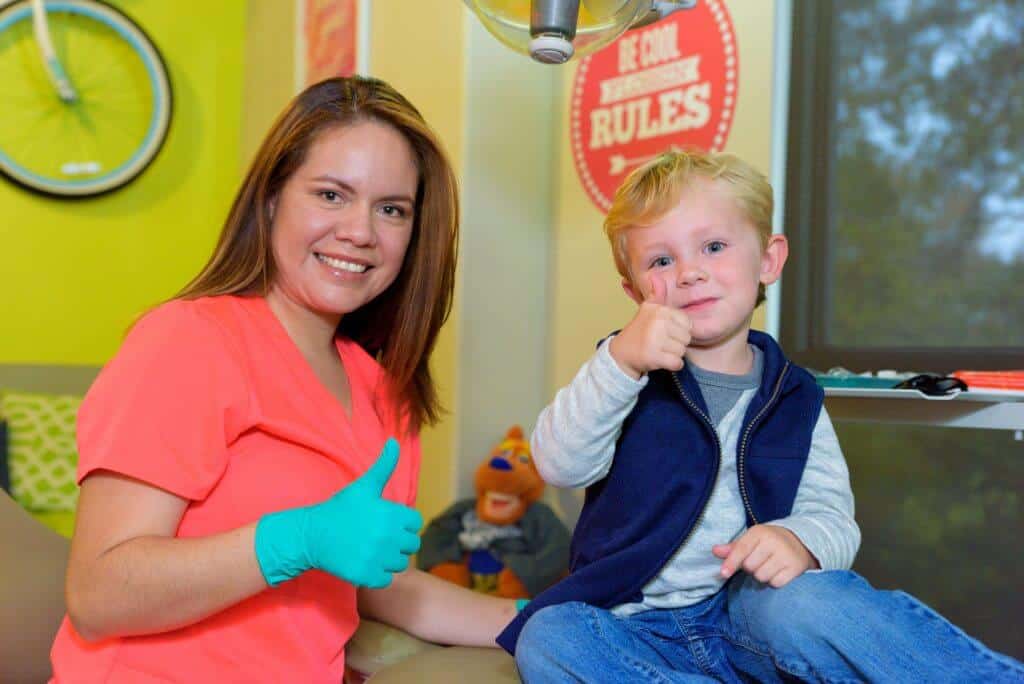 A young patient wearing blue jeans a gray sweatshirt and a blue open zippered vest signaling holding his right thumb up sitting on a treatment bed and a smiling a team members of Hill Country Pediatric Dentistry and orthodontics office wearing orange office uniform and green exam gloves sitting next to the treatment bed on which the blond hair boy is sitting, is also holding her right thumb up the view of the yellow bright wall behind her showing part of the bicycle hanging from the wall